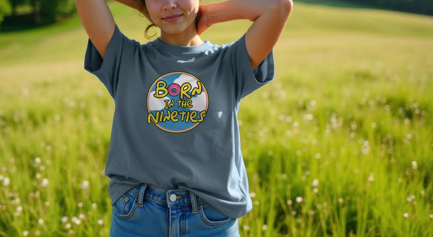 A Woman in a blue Birthday T-shirt with "Born in the Nineties" printed on it stands in a sunny field. Their hands are behind their head, conveying a relaxed mood.