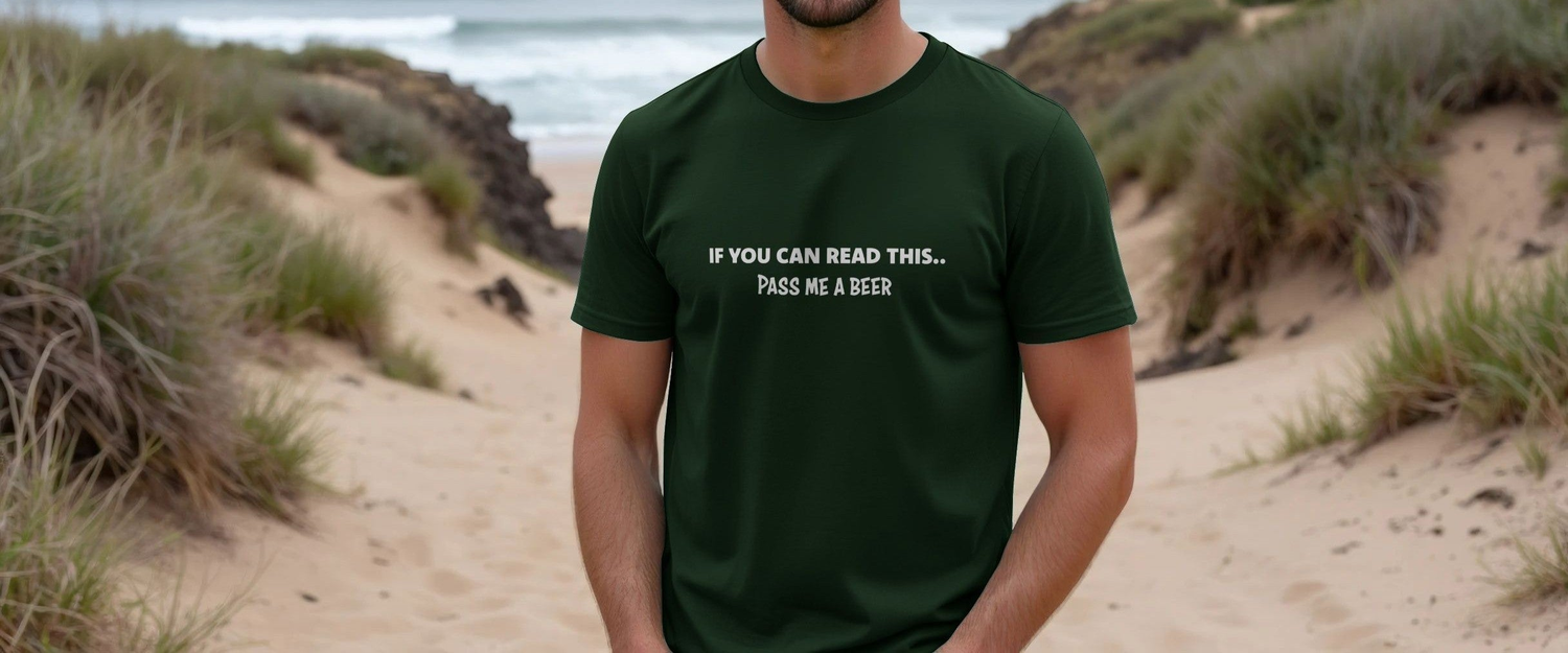 Man in a green Funny Slogan T-shirt with the text “If you can read this.. pass me a beer,” standing on a sandy beach path with grass and ocean waves in the background.