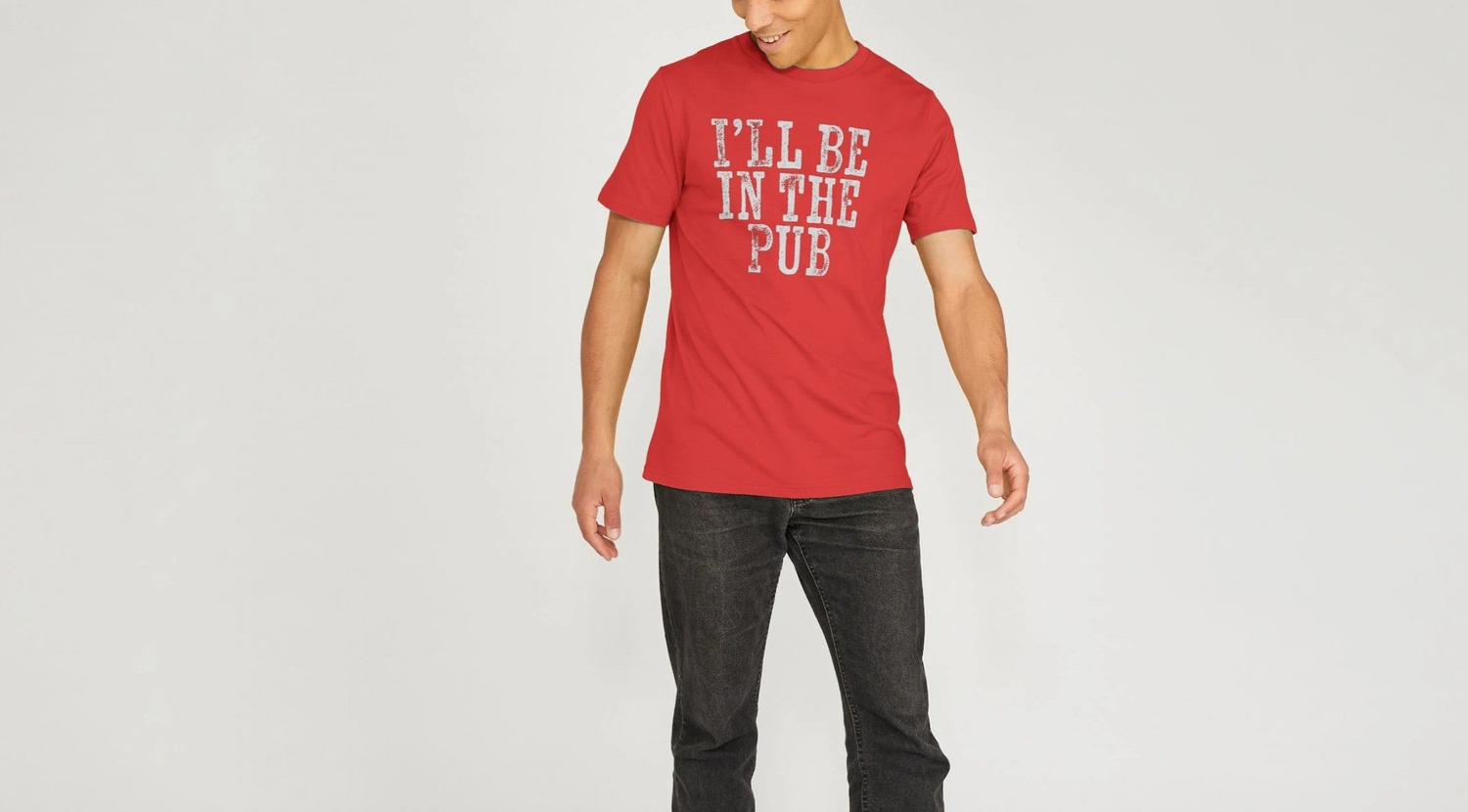Man in a red "I'll be in the Pub" Dad T-shirt with white text saying "I'll Be in the Pub" smiles while looking down. He wears black jeans, standing against a plain background. Casual and playful tone.