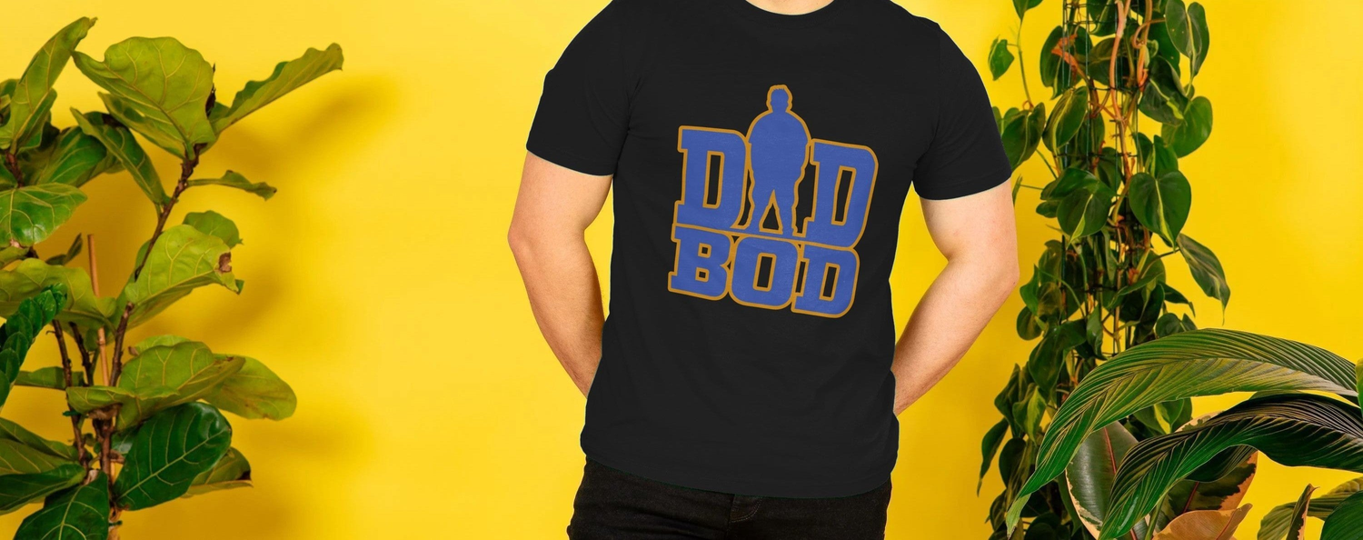 A Dad in a black "Dad Bod" T-shirt stands against a vibrant yellow wall. Green potted plants frame the scene, creating a lively, playful atmosphere.