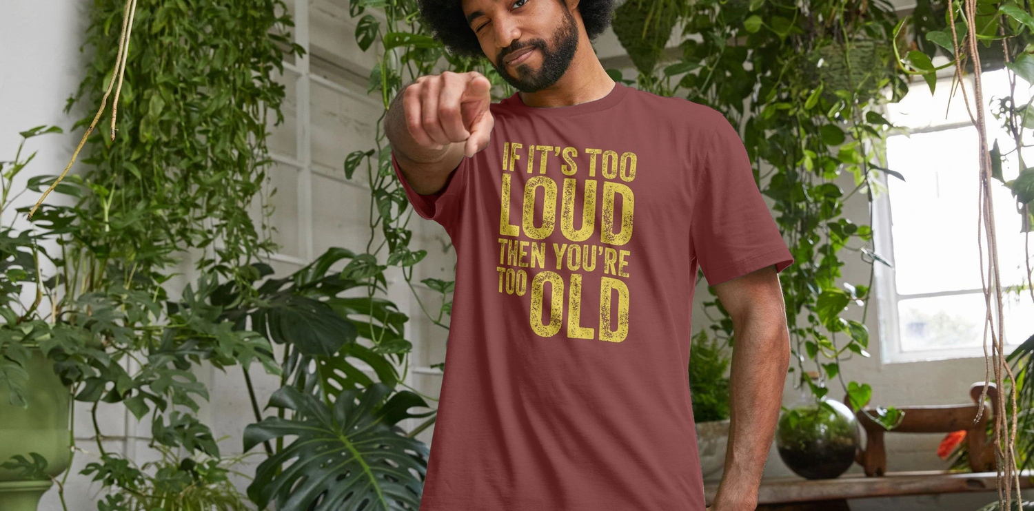 A man with curly hair winks while pointing forward, wearing a red funny slogan t-shirt saying, "If it's too loud then you're too old." He stands amidst lush indoor plants.
