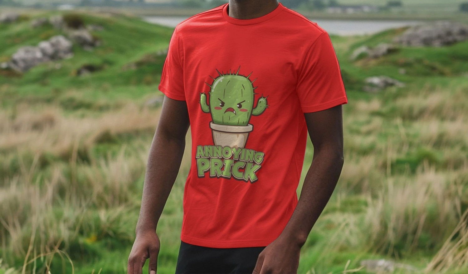 A person wearing a red funny graphic T-shirt featuring an angry cartoon cactus in a pot with the text "Annoying Prick." Background shows a blurred grassy field.