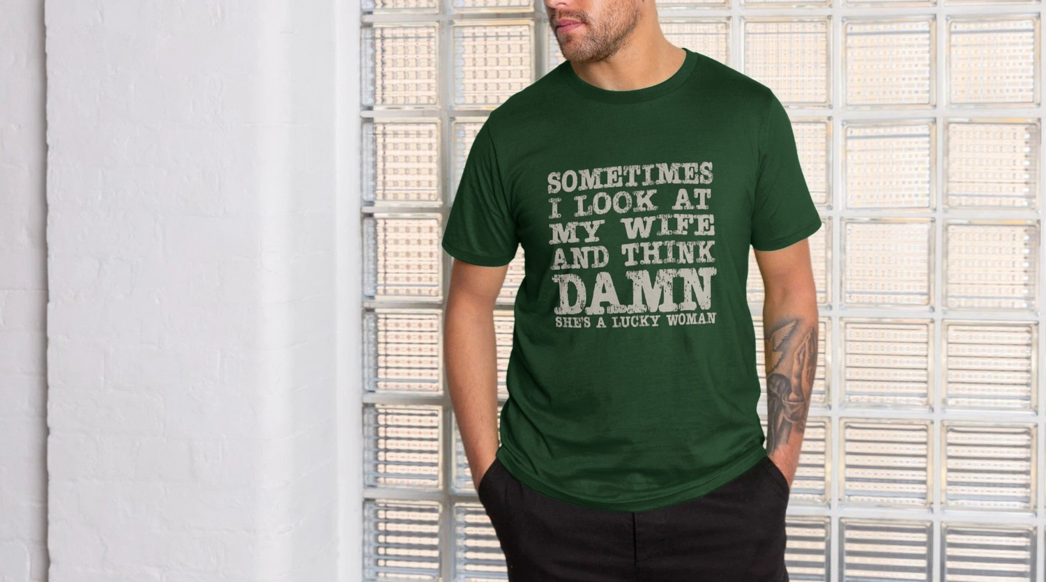 Man in a green Dad Joke T-shirt stands against a textured glass wall. The shirt humorously reads, "Sometimes I look at my wife and think, damn she's a lucky woman."
