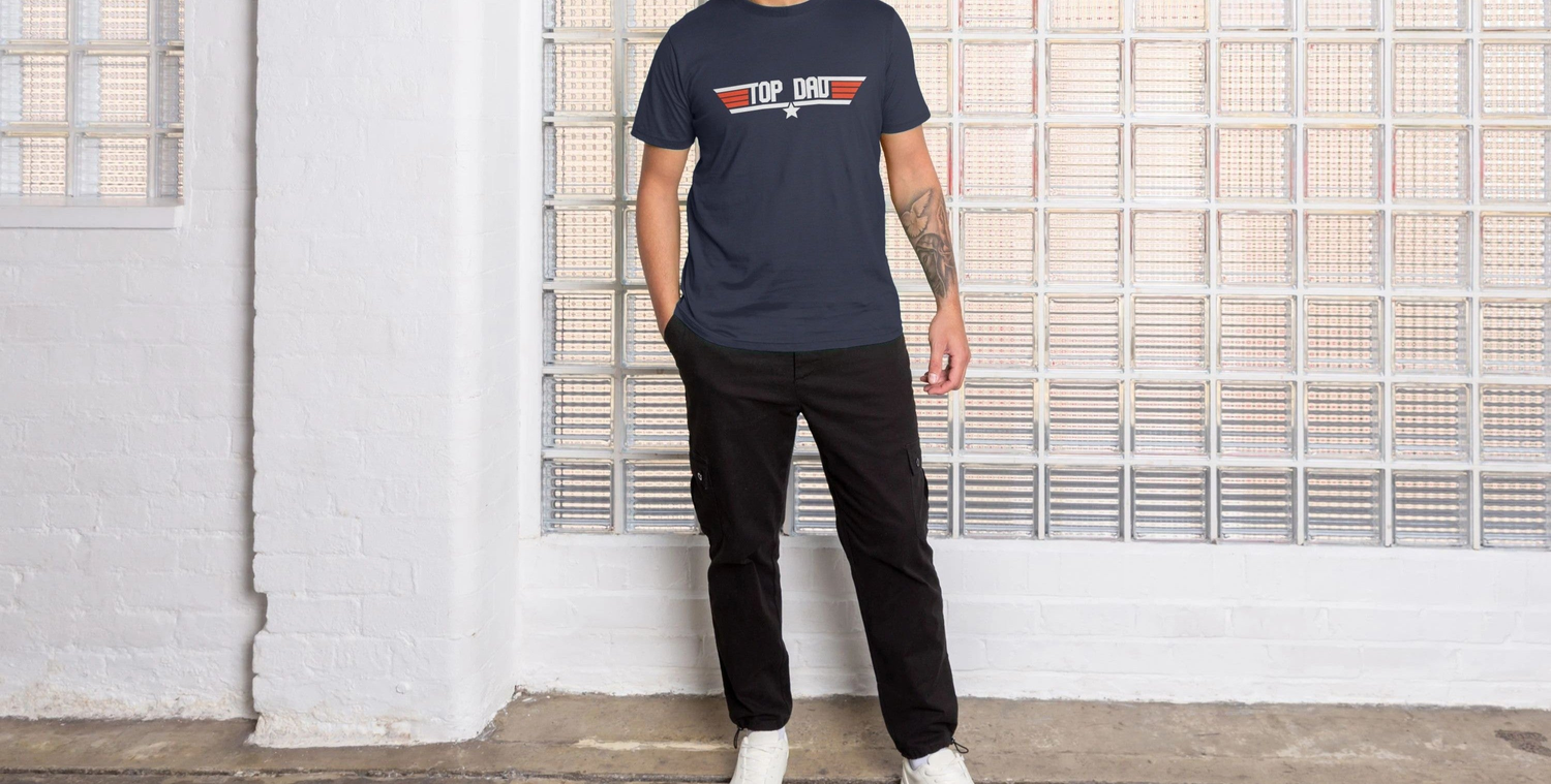 Man wearing a fathers day gift, a navy "Top Dad" T-shirt, black pants, and white shoes stands casually against a glass block wall in a minimalist, bright room.