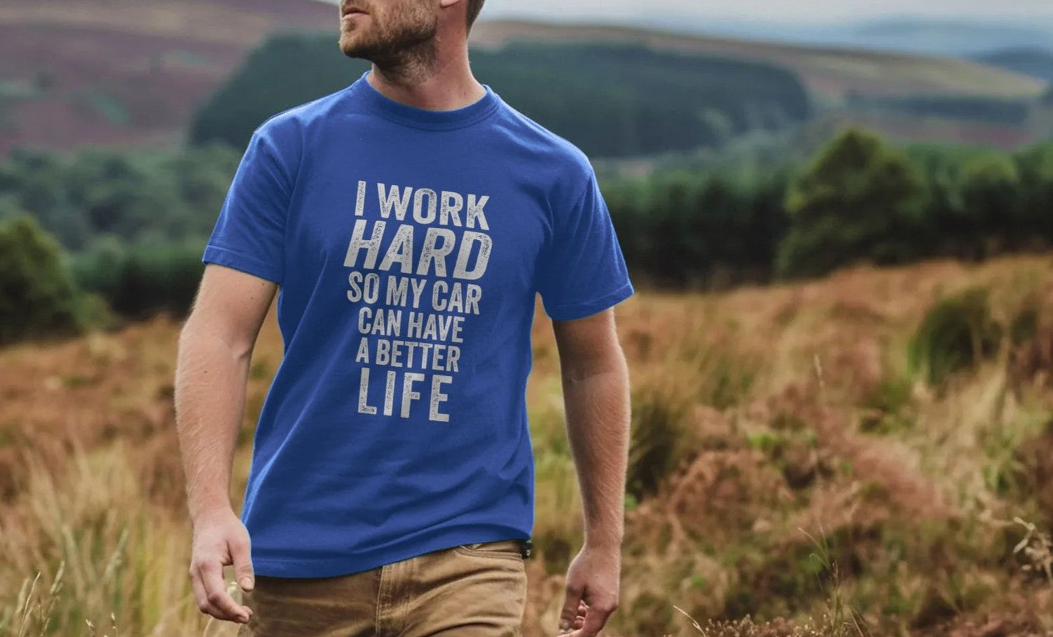 Man in blue Car T-shirt with slogan "I work hard so my car can have a better life" walks through a scenic, grassy landscape. Calm and humorous tone.
