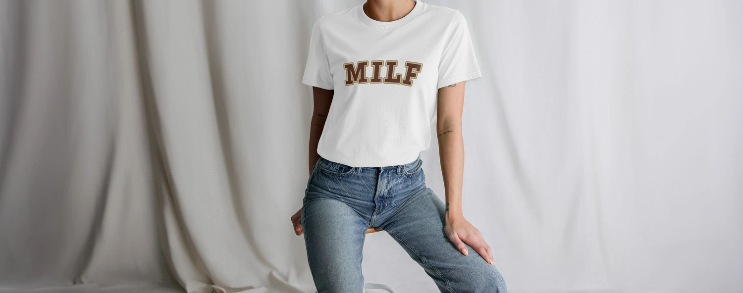 A Mum wearing a white Mum T-shirt with "MILF" in bold brown letters and blue jeans, posing against a gray draped background, exuding a casual vibe.