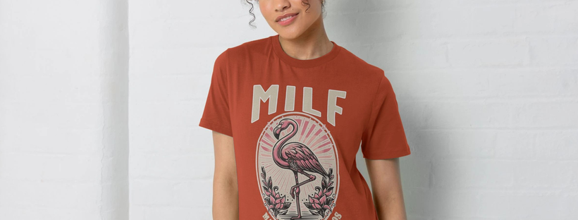 A Woman with curly hair wears a rust-colored Funny Women's t-shirt featuring a flamingo illustration and large text saying MILF (Man I Love Flamingo's) . They stand against a white brick wall, smiling slightly.
