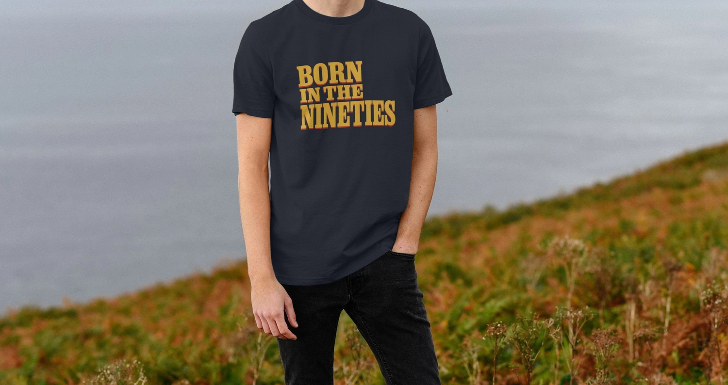 Man in a "Born in the Nineties" Birthday t-shirt stands on a grassy hilltop overlooking the ocean. The scene conveys a serene and reflective mood.