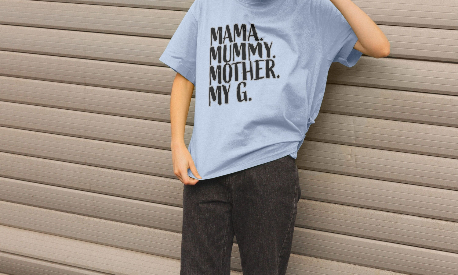 A Mum in a light blue Mum T-shirt with black text reading "Mama. Mummy. Mother. My G." leans against a beige, horizontally-striped wall, creating a relaxed tone.