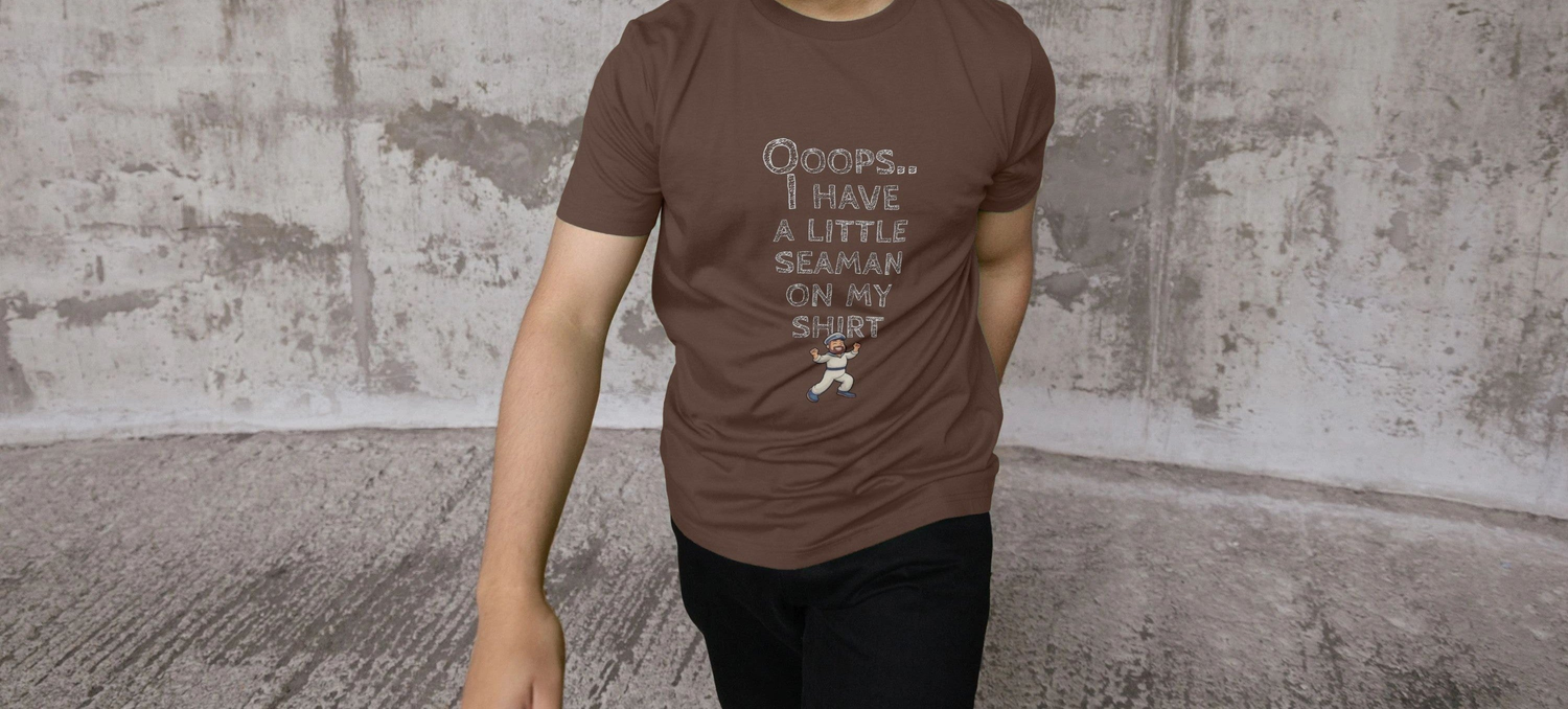 A Man with short black hair walks in front of a textured concrete wall, wearing a brown Funny Rude T-shirt that says "Oops... I have a little seaman on my shirt."