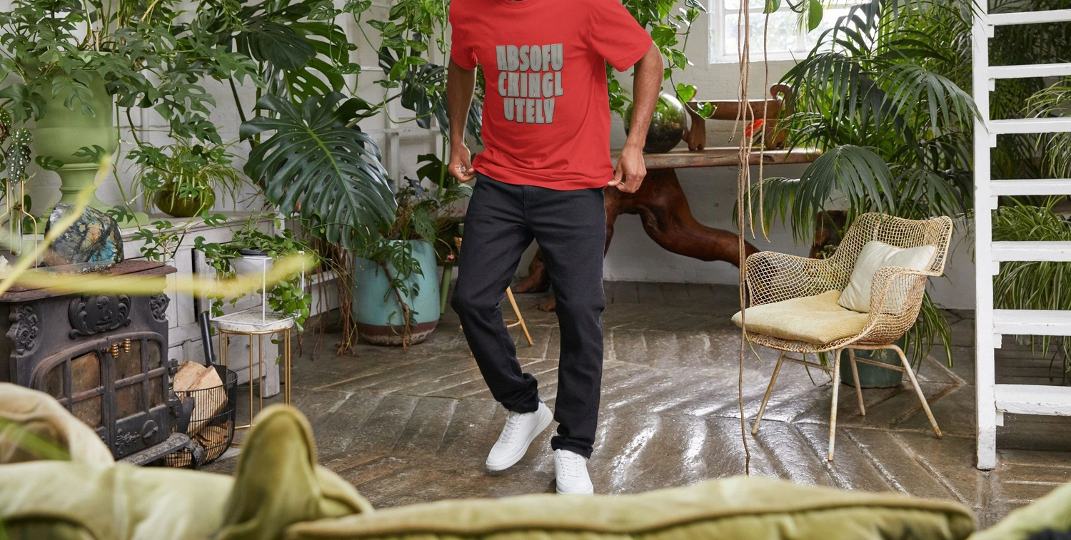 Man in a red funny sarcastic T-shirt with the words "ABSOFU CKING LUTELY," stands joyfully in a lush room with hanging plants, conveying a relaxed, vibrant vibe.