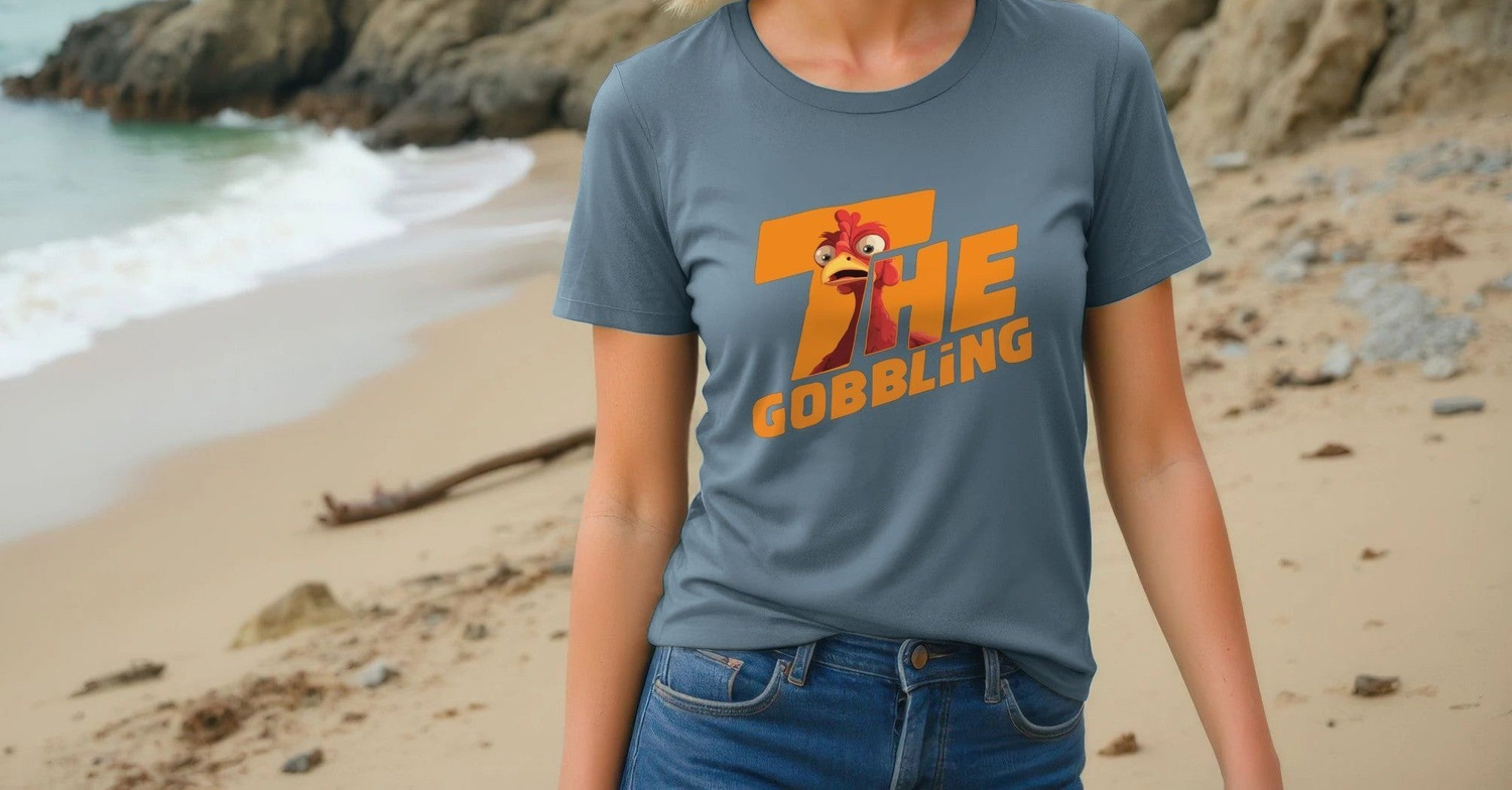 A Woman in a gray Christmas T-shirt with a cartoon turkey and "The Gobbling" in bold, vibrant text stands on a sandy beach with rocky cliffs and gentle waves.