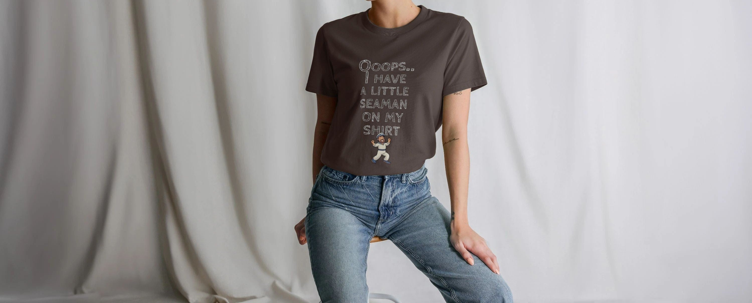 A Woman in jeans sitting casually, wearing a dark Womens Rude t-shirt with humorous text "Whoops I have a little seaman on my shirt" and a cartoon sailor. The backdrop is a soft, neutral-toned fabric.