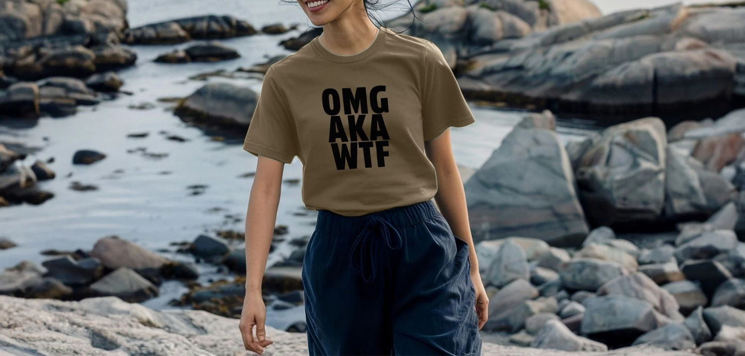 A woman wearing a beige Sarcastic T-shirt with bold black text "OMG AKA WTF" stands smiling by a rocky shoreline, creating a playful and relaxed vibe.