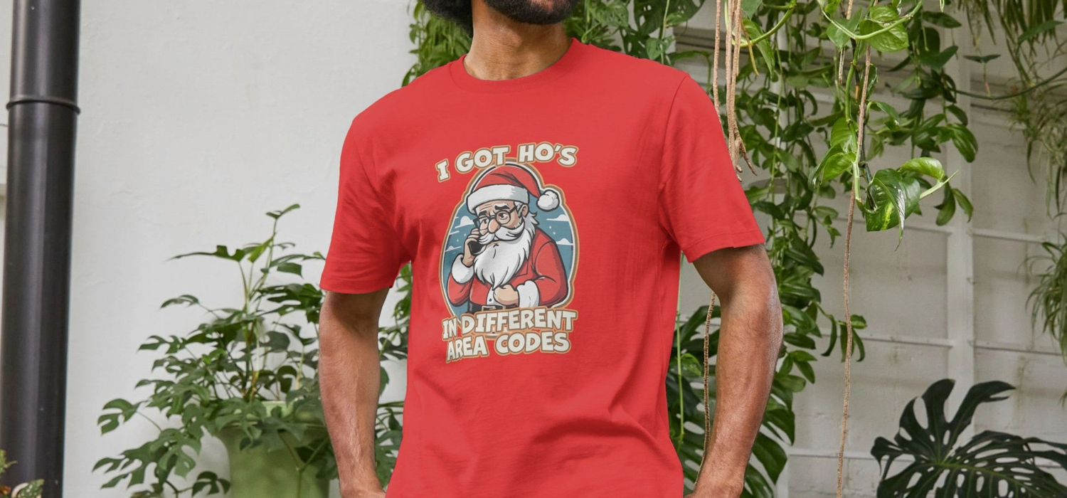 Man in a red Christmas T-shirt featuring a Santa graphic with humorous text stands in a lush setting. The design adds a playful, festive tone.