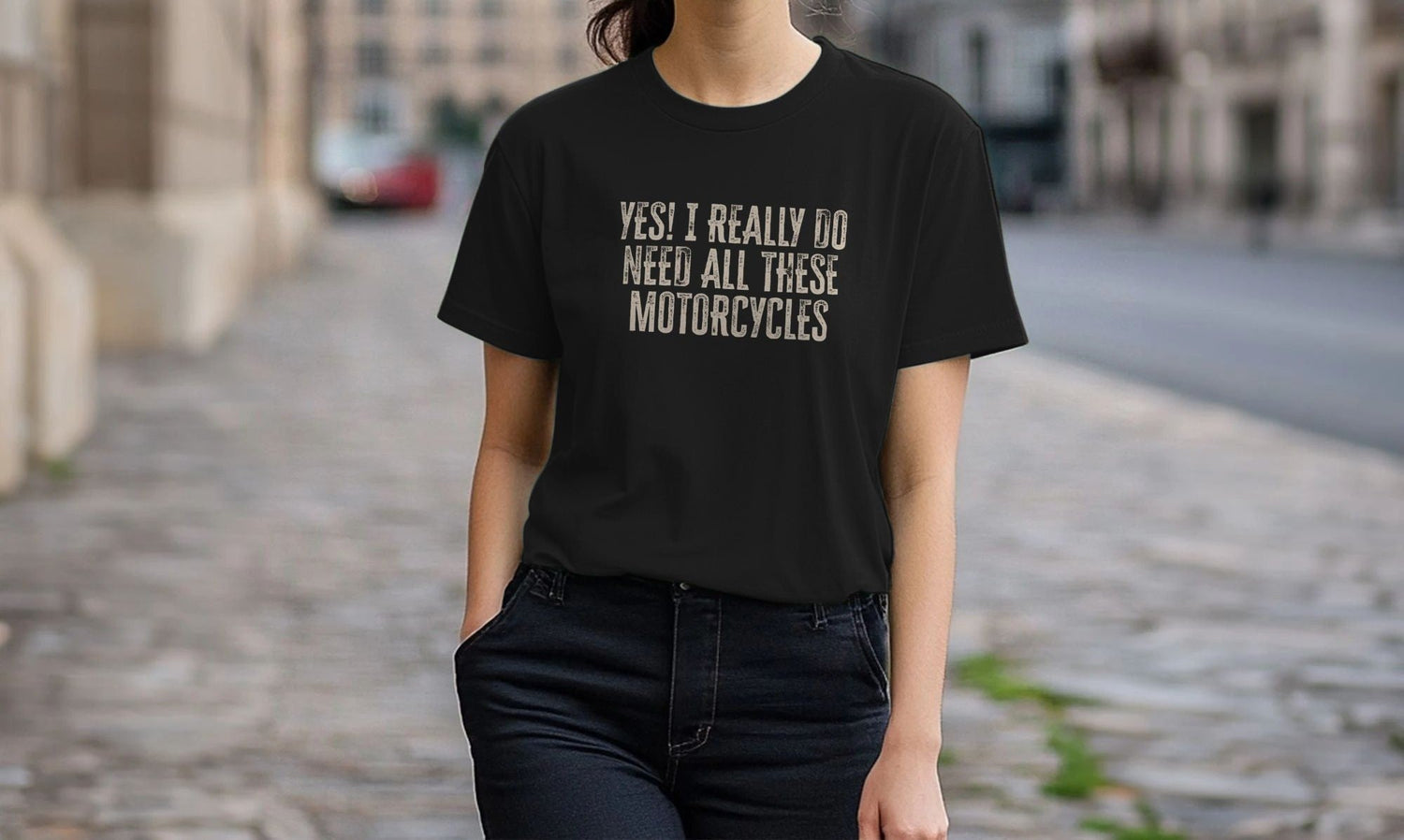 A Woman wearing a black Motorcycle T-shirt with the text "Yes! I really do need all these motorcycles" stands casually on a cobblestone street, conveying a relaxed and humorous tone.