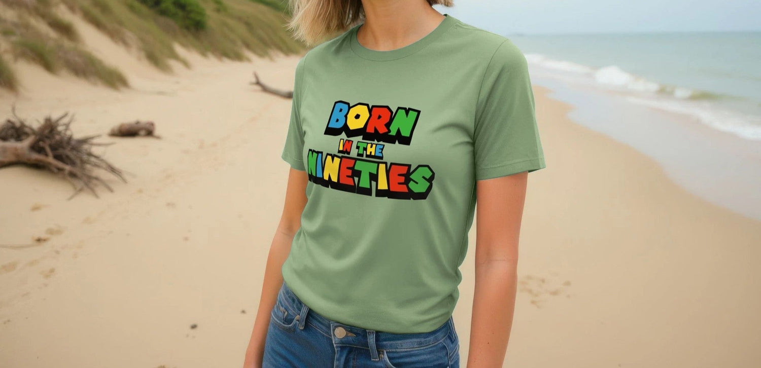A woman stands on a sandy beach wearing a green "Born in the Nineties" Birthday T-shirt. The background features grassy dunes and a calm sea, creating a relaxed vibe.