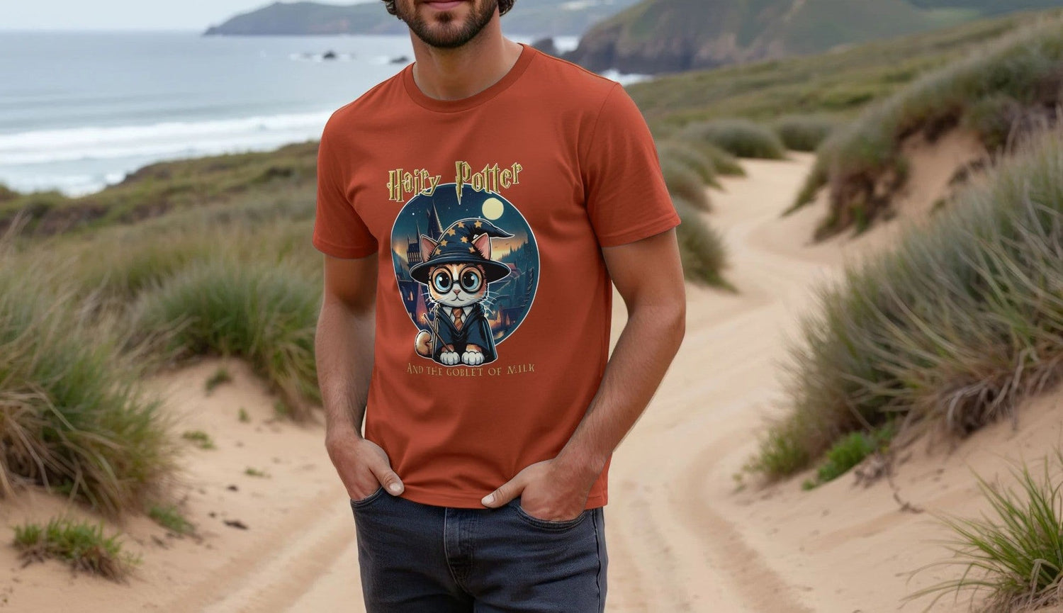 Man in an orange Geek t-shirt with a whimsical cat illustration, "Hairy Potter and the Goblet of Milk," standing on a sandy path with ocean and hills in the background.