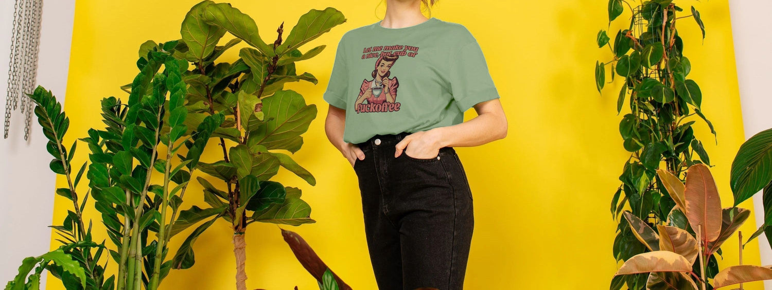 A Women stands against a bright yellow backdrop, surrounded by lush green plants. They wear a pastel green Retro Graphic T-shirt with bold text and graphics, exuding a playful, vibrant vibe.
