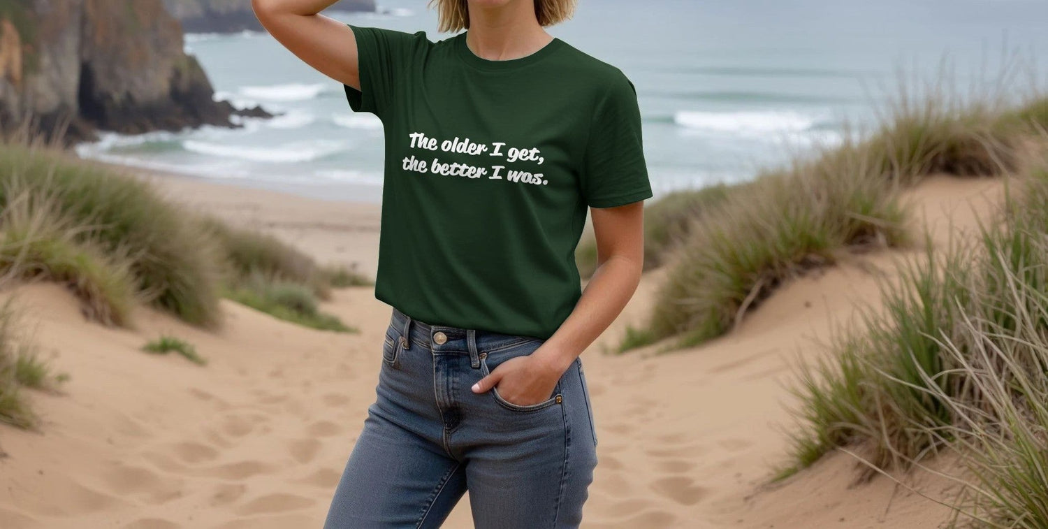 A woman in a green Retirement Gift T-shirt with the text "The older I get, the better I was" stands on a sandy beach. Dunes and ocean waves add a serene backdrop.