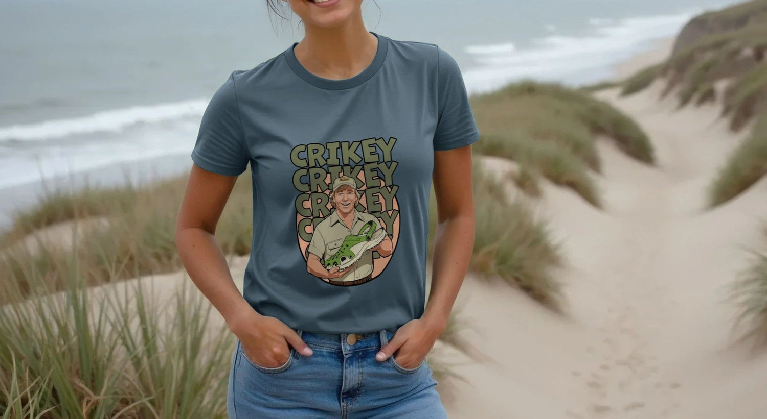 Woman at the beach in a blue Movie and TV T-shirt with a cartoon of a man that looks like Steve Irwin holding a crocodile, repeating the word "Crikey." Sandy dunes in the background.