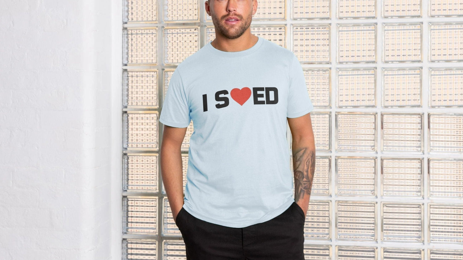 A man in a light blue funny offensive t-shirt with "I ♥ ED" stands against a textured glass brick wall. His hands in pockets, he expresses a relaxed and casual vibe.