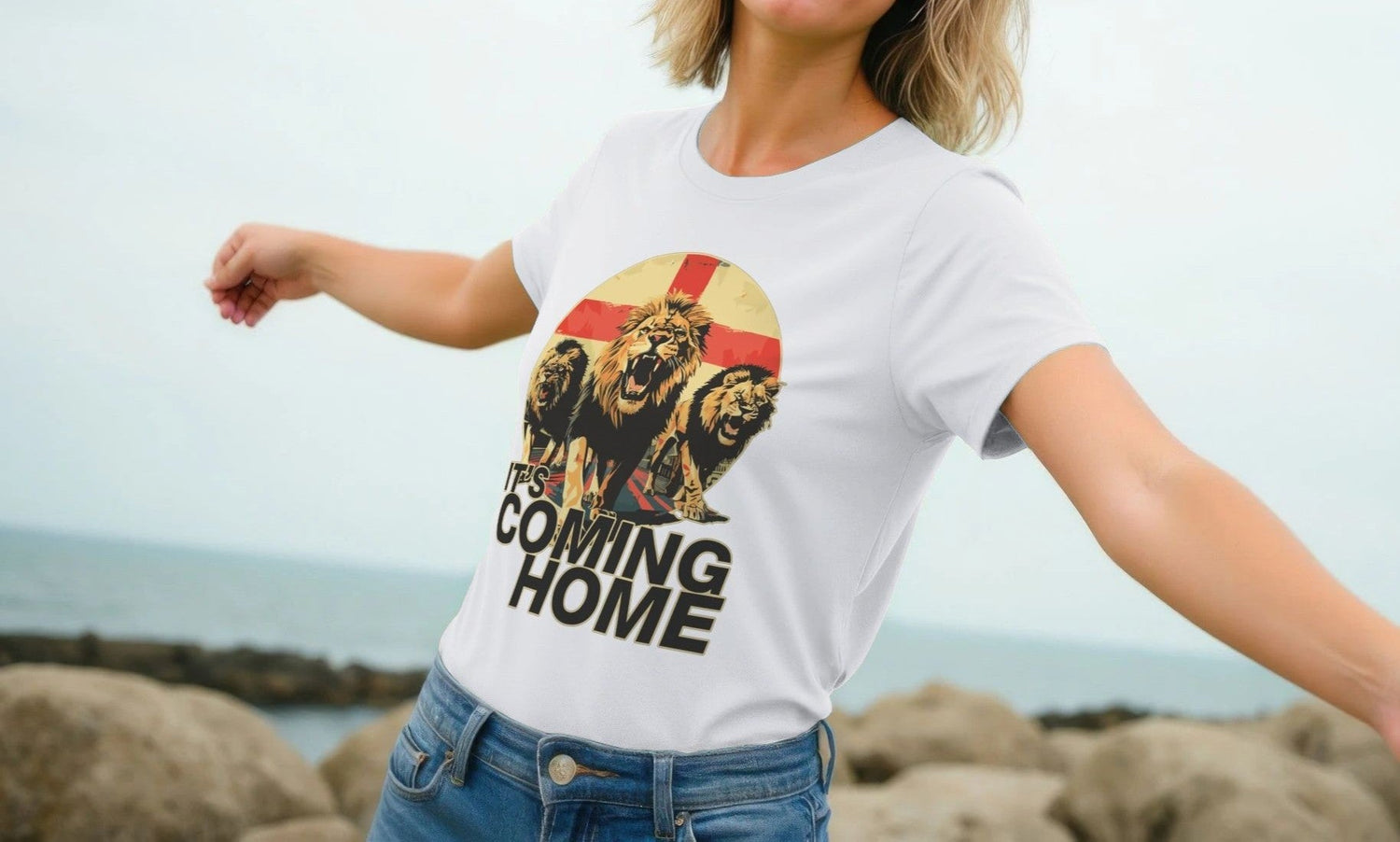 A Woman stands near a rocky shore, arms spread wide, wearing a white Women's Sports T-shirt featuring three roaring lions and the words “It's Coming Home” on an England flag.