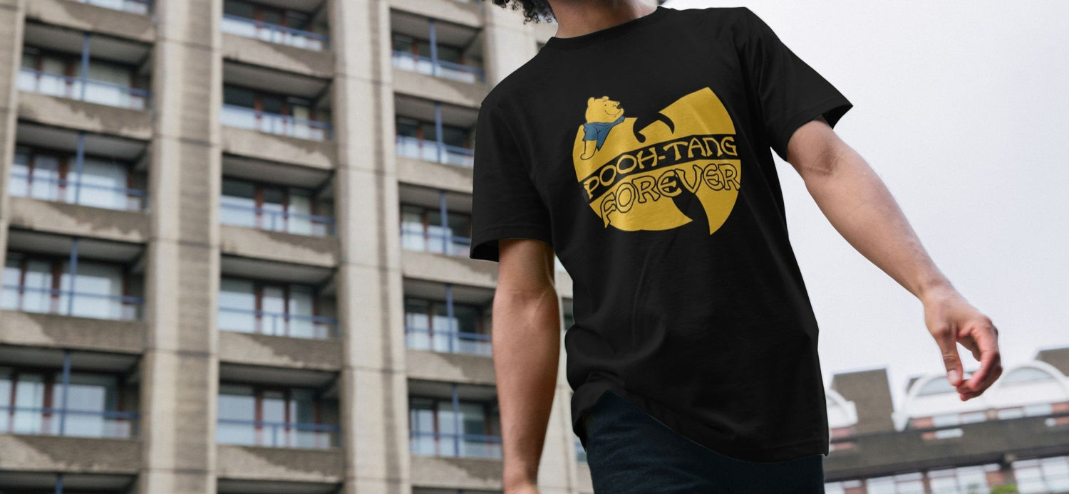 A Man wearing a black Festival t-shirt with a yellow graphic featuring a bear and the Logo text "Pooh-Tang Forever" walks in front of a concrete building. Urban, casual vibe.