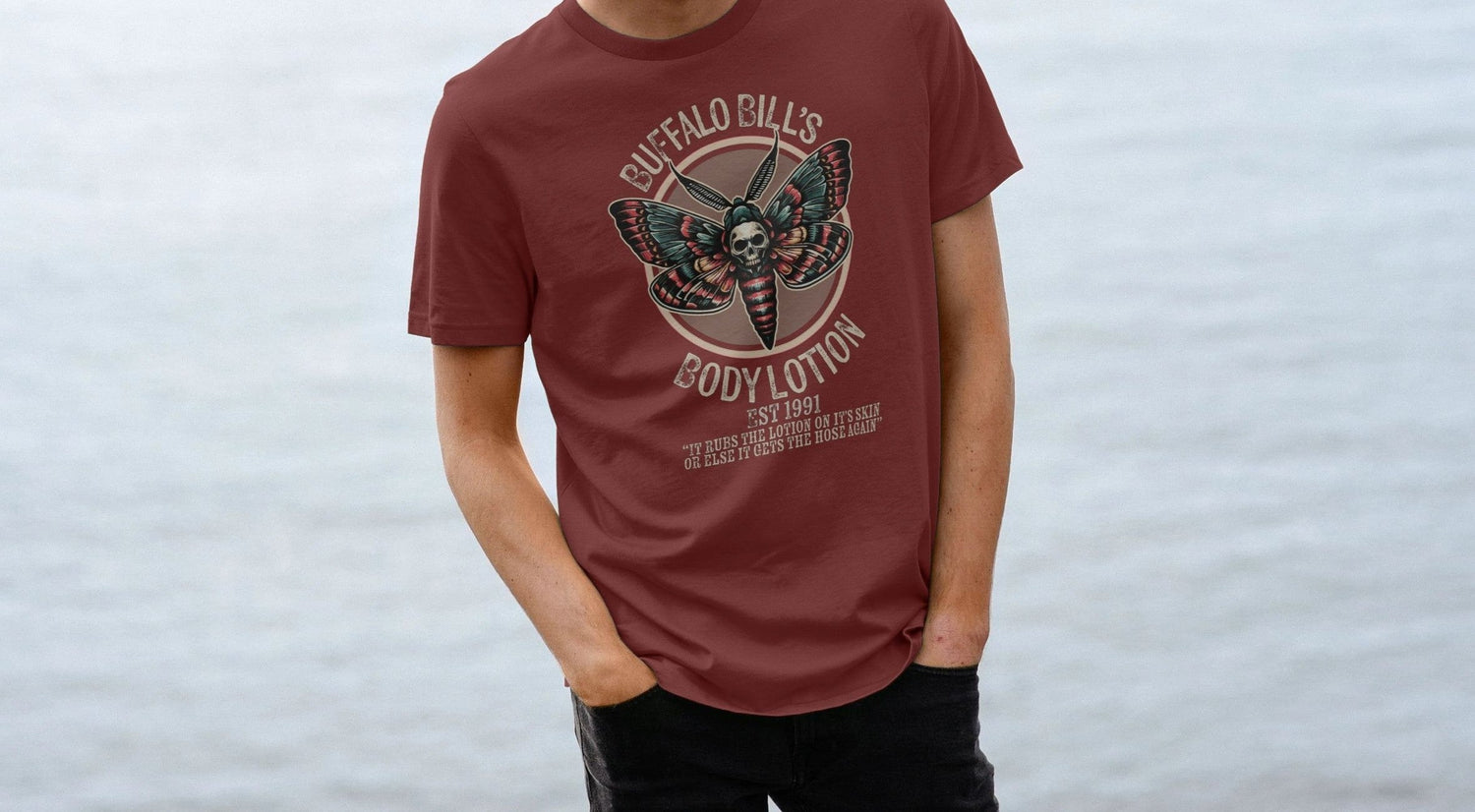 Young man in a maroon Horror T-shirt stands by calm water. The shirt features a design with a moth and skull, reading "Buffalo Bill's Body Lotion." Casual and pensive mood.