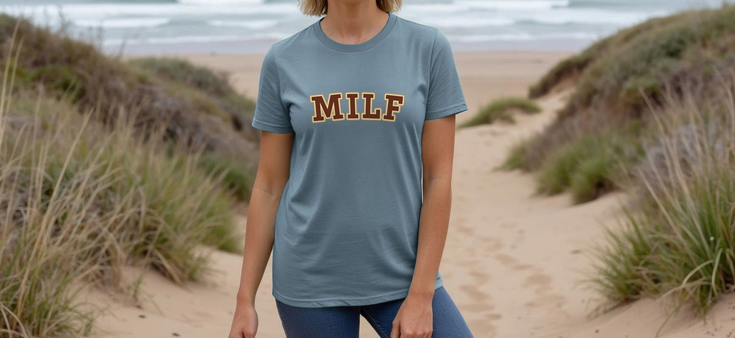 A Woman stands on a sandy path between grassy dunes, facing the camera with a calm expression. They wear a blue Hen Party T-shirt with the word "MILF" in large letters. The ocean and waves are visible in the background under a cloudy sky.