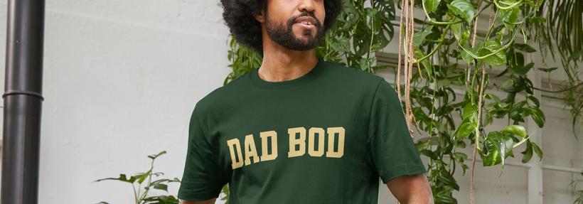 Man with curly hair and a beard wearing a Fathers Day Gift of a green "DAD BOD" t-shirt stands in a garden-like indoor space. The atmosphere is relaxed and natural.