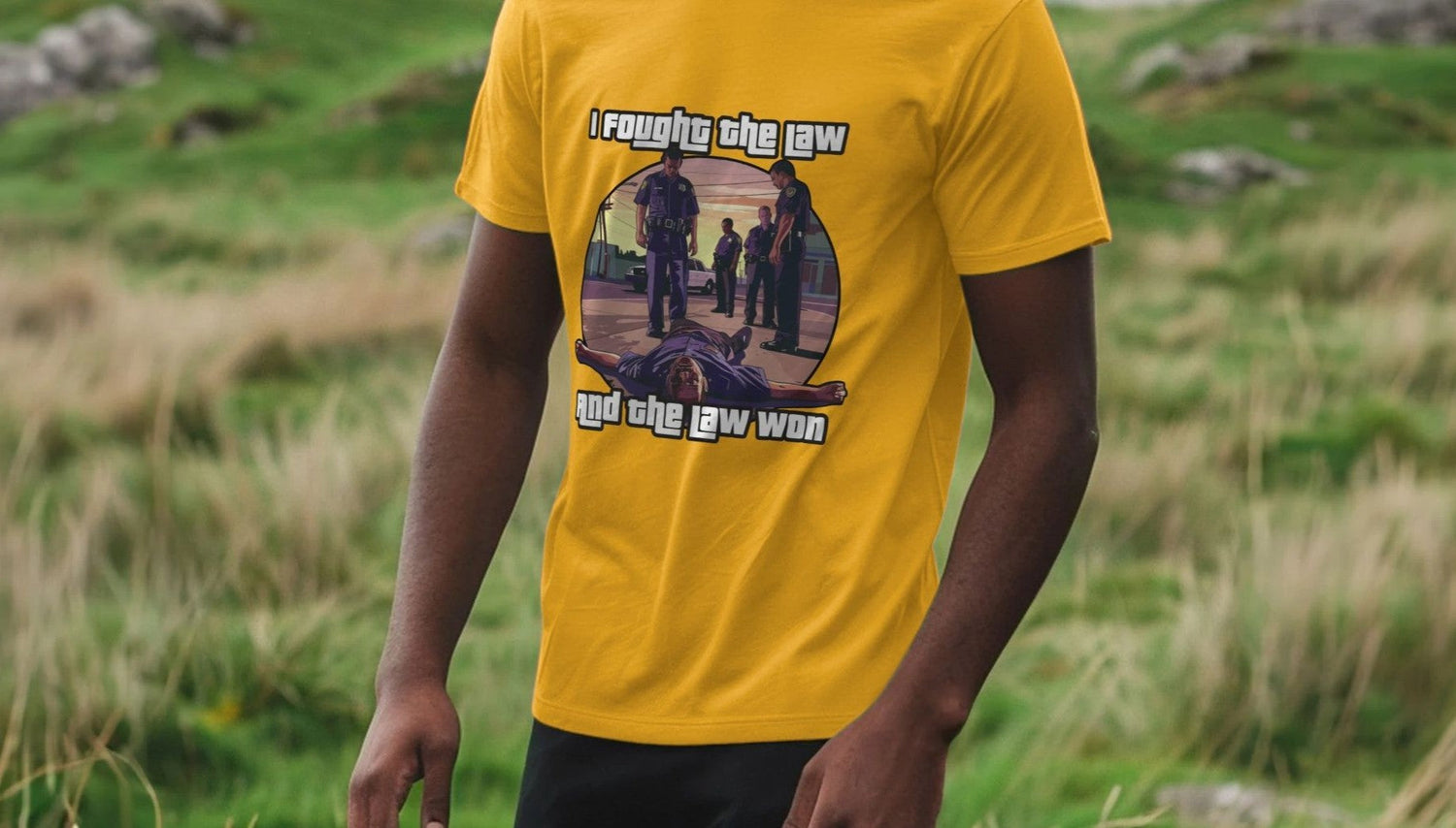 Man wearing a yellow Gaming T-shirt with an illustration of police officers and a person on the ground. Text reads, "I fought the law and the law won." Background shows lush, green grass.