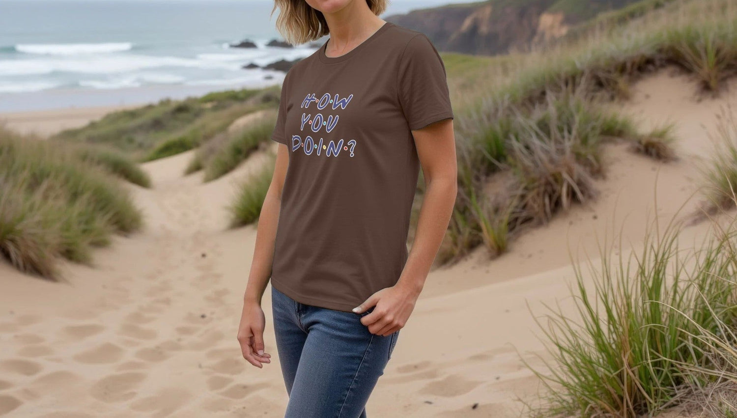 Woman wearing a brown Geek T-shirt with "How You Doin'?" in a Friends style font, stands on a sandy beach path, surrounded by grass, with waves and cliffs in the background.