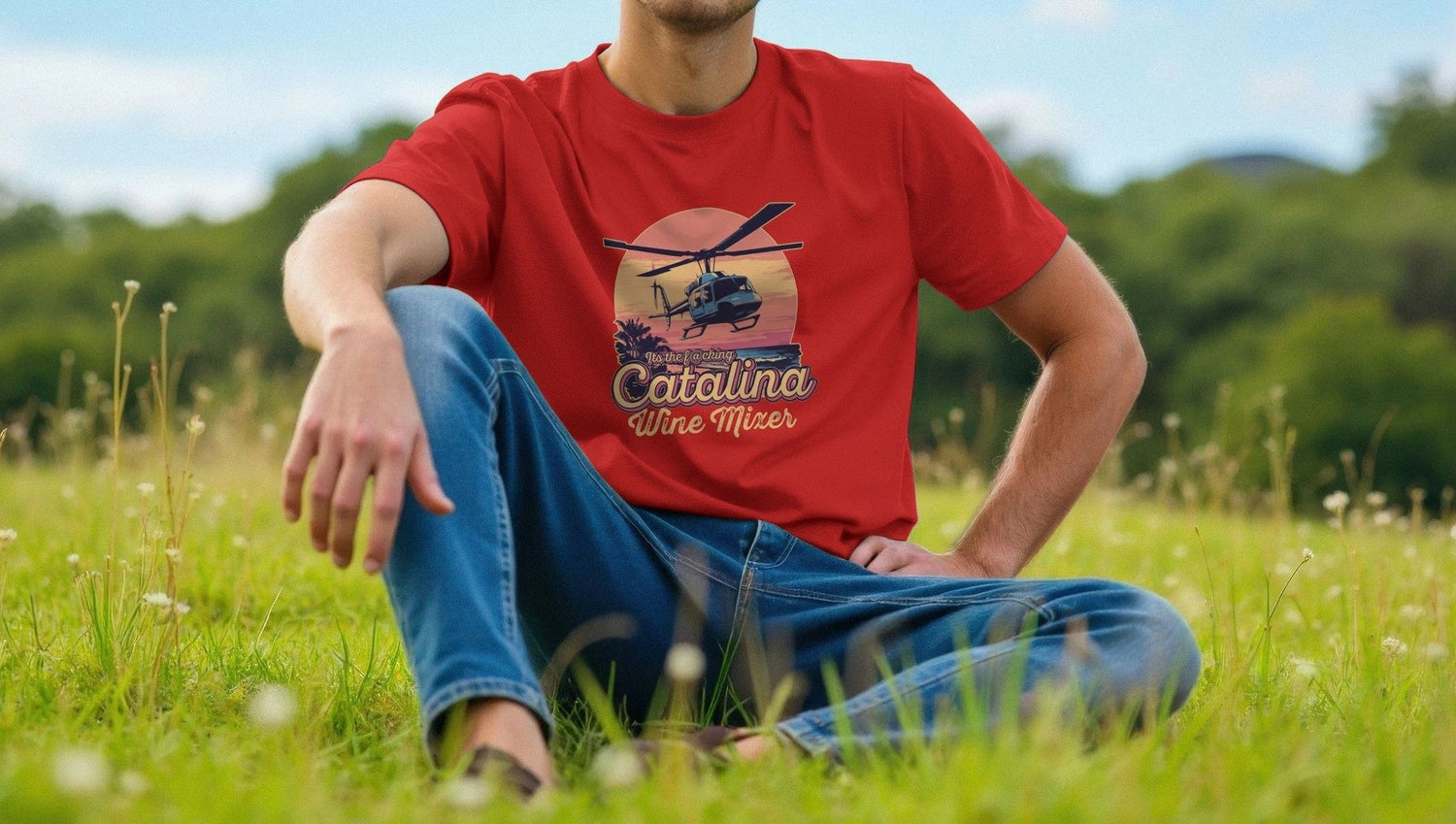A Man sits on grass wearing a red Movie t-shirt with "Catalina Wine Mixer" and a helicopter design, inspired by "Step Brothers". The background features green trees and a clear sky.