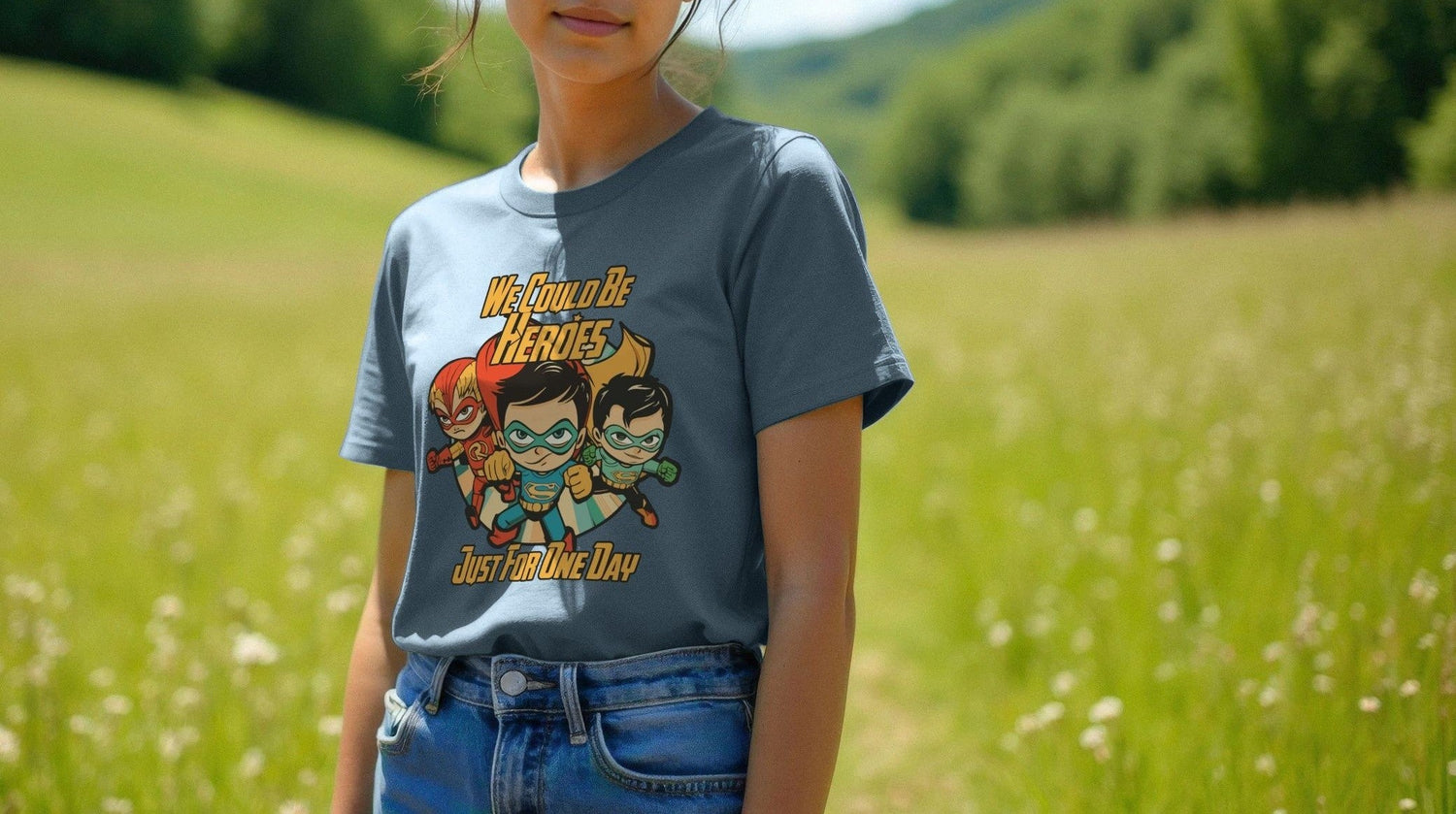 Young woman in a field wearing a Women's Festival T-shirt with cartoon heroes and the text "We Could Be Heroes Just For One Day." Sunny, peaceful outdoor setting.