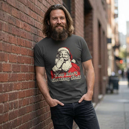 Long-haired man in khaki You Ain’t Getting Shit Santa tee against brick wall, fun festive xmas gift