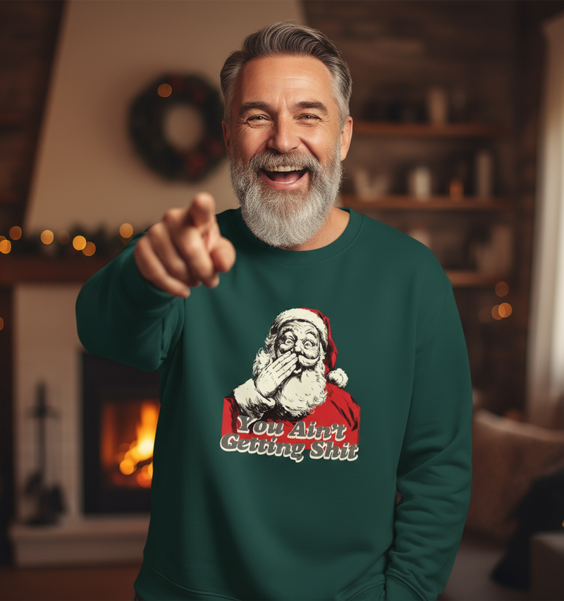Man wearing a green Rude Santa sweater with a Santa Claus graphic and text, pointing forward in a cozy room.
