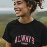 A woman stands outdoors in a scenic landscape, wearing a black t-shirt with pink lettering that reads 