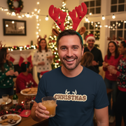 Man wearing reindeer antlers and a 'Christmas' shirt, holding a drink, in a festive indoor setting with people and decorations.