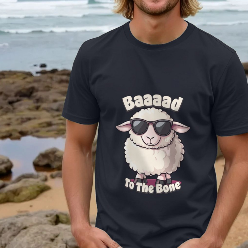 A person stands on a rocky beach wearing a black funny animal T-shirt featuring a cartoon sheep in sunglasses with the text "Baaaad to the Bone," creating a playful vibe.