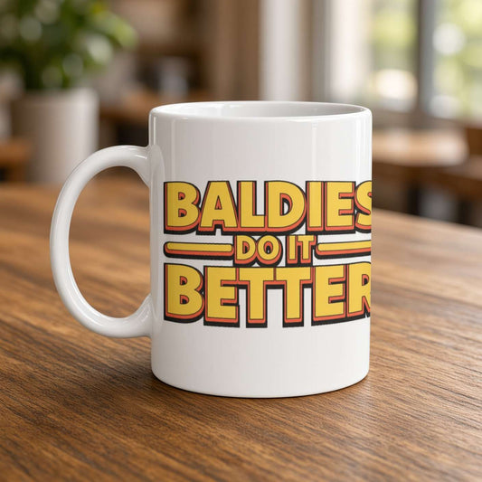 A white coffee mug on a wooden table reads “Baldies do it better” in bold red-and-yellow lettering, creating a playful, upbeat mood.