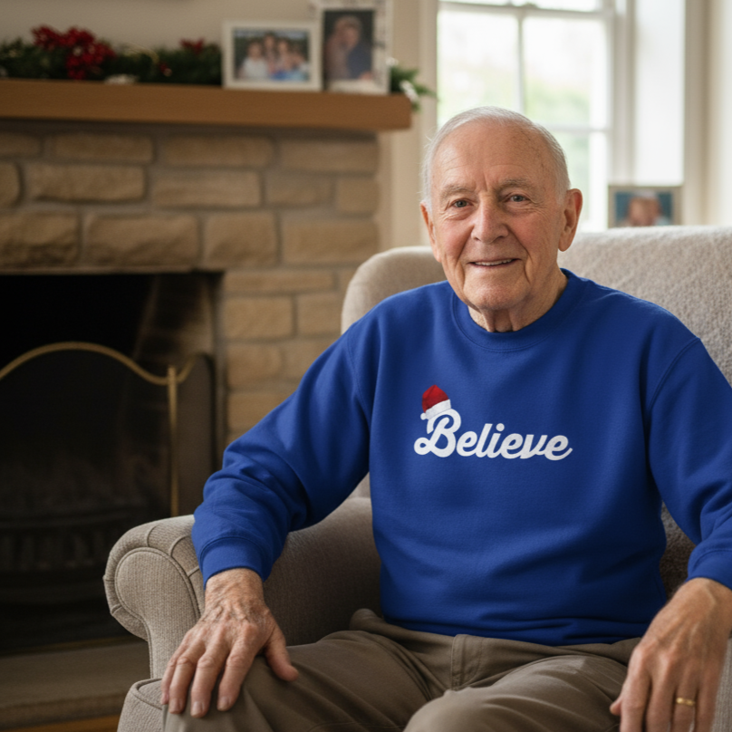 Man wearing a blue Christmas sweatshirt with 'Believe' on it, sitting in a living room.