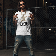 Man wearing a white funny jesus t-shirt with graphic design and text, standing in front of a chain-link fence.