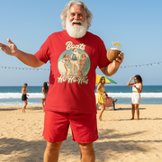 Santa in red funny Christmas shirt and shorts holding a coconut on a beach with people in the background