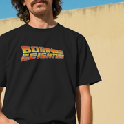 Person wearing a black birthday t-shirt with 'Born in the Eighties' slogan in a back to the future style font, against a beige wall.