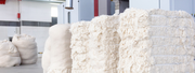 Stacks of compressed wool bundles in a bright, industrial setting. The wool appears fluffy and soft, giving a sense of abundance and texture.
