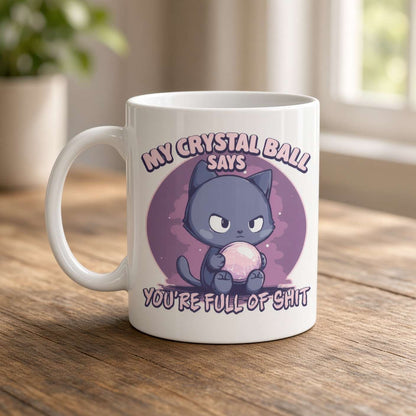 A white mug on a wooden table features a grumpy cartoon cat holding a pink crystal ball, with the joke text, “My crystal ball says you’re full of shit.” The tone is playful and irreverent.