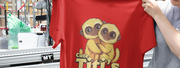 A person holds a red T-shirt featuring two cartoon titi monkeys hugging, with the text 