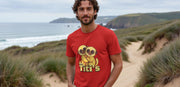 A man stands on a sandy, grassy coastal path wearing a red funny animal T-shirt with cute cartoon titi monkeys hugging. The background shows a cloudy sky and ocean.