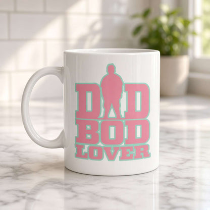 A white ceramic mug on a marble counter reads “DAD BOD LOVER” in bold pink and teal lettering, with a dad silhouette; warm, cheerful kitchen setting.